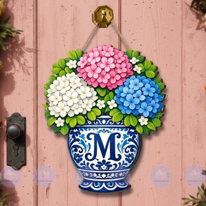 May include: A wooden door sign featuring a blue and white vase with the letter "M" and a bouquet of pink, white, and blue hydrangeas. The sign is hanging from a gold-colored knob with a rope.