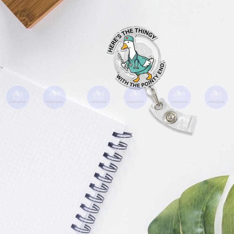 May include: A clear badge reel featuring a cartoon duck wearing a teal medical coat and holding a scalpel. The text on the badge reel reads "Here's the thingy with the pointy end."