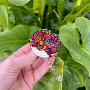 Könnte beinhalten: Ein weißer, einziehbarer Ausweishalter mit einem farbenfrohen floralen Gehirndesign. Der Ausweishalter ist an einem durchsichtigen Plastikband befestigt.