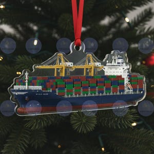 Op de afbeelding: Transparante acryl kerstversiering in de vorm van een containerschip. Het blauwe schip heeft rode en witte details en is geladen met kleurrijke zeecontainers. Twee gele kranen zijn zichtbaar. Een rood lint is bevestigd om op te hangen.