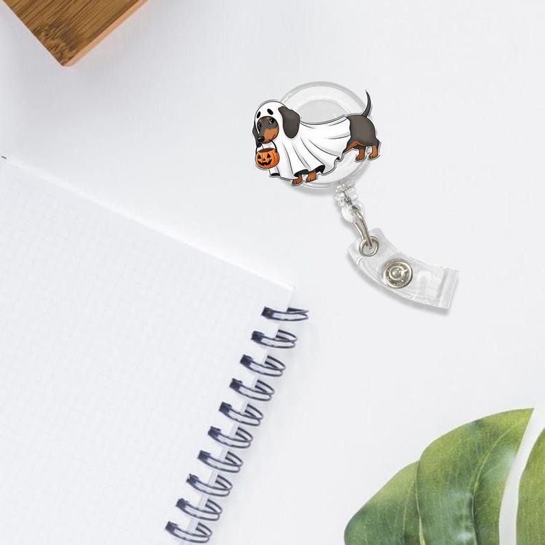 Puede incluir: Un portatarjetas retr&aacute;ctil con tem&aacute;tica de Halloween con un perro salchicha disfrazado de fantasma, sosteniendo una calabaza. El dise&ntilde;o est&aacute; sobre un carrete de pl&aacute;stico transparente con un clip met&aacute;lico. El fondo es blanco.