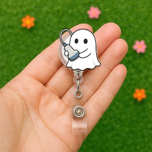 Puede incluir: Un portatarjetas en forma de fantasma blanco con un diseño de dibujos animados que sostiene una sonda de ultrasonido médico. El portatarjetas tiene una correa de plástico transparente y un clip de metal. El fondo es verde con flores de colores.