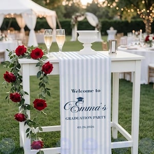 May include: A white table decorated for a graduation party. A blue and white striped runner displays the text "Welcome to Emma's Graduation Party" with the date 05.26.2026. Red roses and greenery cascade down the side. Two champagne flutes and a white vase are on the table.