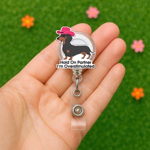 May include: A clear retractable badge reel featuring a cartoon dachshund wearing a pink cowboy hat. The dachshund is black and brown with a white background. The text "Hold On Partner I'm Overstimulated" is below the dog.