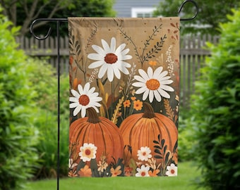 May include: A garden flag featuring a fall theme with two orange pumpkins and large white daisies with brown centers. The background is a mix of brown and green with additional floral elements. The flag is hanging on a black metal stand.