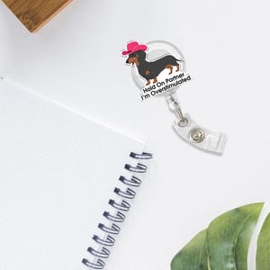 May include: A clear badge reel with a cartoon dachshund wearing a pink cowboy hat. The text "Hold On Partner I'm Overstimulated" is printed below the dog. The reel has a clear plastic clip.