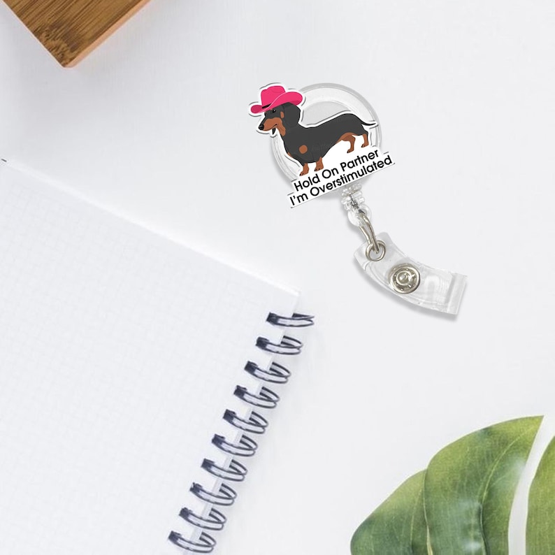 May include: A clear badge reel with a cartoon dachshund wearing a pink cowboy hat. The text "Hold On Partner I'm Overstimulated" is printed below the dog. The reel has a clear plastic clip.