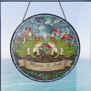 May include: A stained glass window hanging with a circular frame. The design features two green frogs with red eyes, sitting on a branch with mushrooms and flowers. The text "Megan & Josh 2.15.2022" is written on a banner below the frogs.