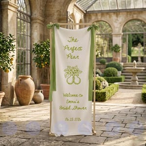 May include: A cream-colored banner with green text and accents, displayed on a wooden frame. The banner reads "The Perfect Pear" and "Welcome to Emma's Bridal Shower" with the date 06.25.2026. Green ribbons are tied at the top.