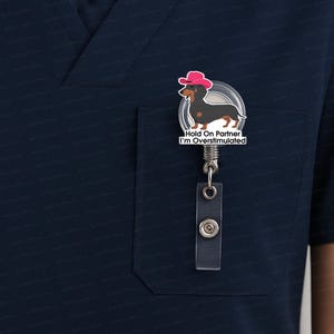 May include: A retractable badge reel featuring a cartoon dachshund wearing a pink cowboy hat. The dachshund is black and brown. The badge reel has the text "Hold On Partner I'm Overstimulated". The reel is attached to a clear plastic holder.