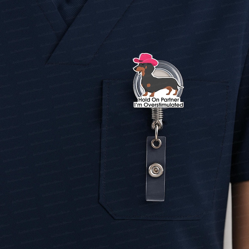 May include: A retractable badge reel featuring a cartoon dachshund wearing a pink cowboy hat. The dachshund is black and brown. The badge reel has the text "Hold On Partner I'm Overstimulated". The reel is attached to a clear plastic holder.