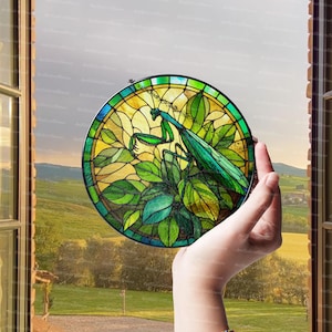 May include: A round stained glass window featuring a green praying mantis among green leaves, set against a yellow and green background. The window is held up in front of a scenic outdoor view.