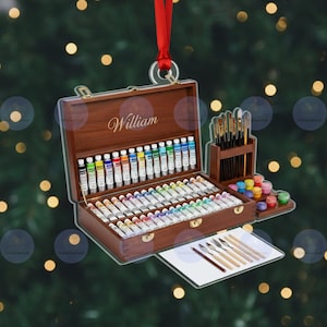 Könnte beinhalten: Ein hölzernes Künstler-Malset-Ornament mit dem Namen "William" in goldener Schrift. Das Set enthält Farbtuben, Pinsel, Farbpaletten und Malmesser. Ein rotes Band ist zum Aufhängen befestigt.