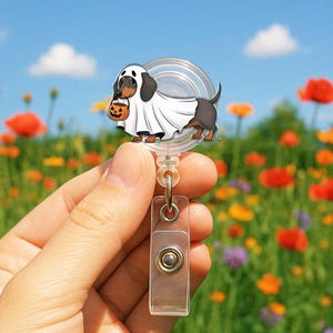 Puede incluir: Un portatarjetas retr&aacute;ctil con tem&aacute;tica de Halloween con un perro salchicha disfrazado de fantasma, sosteniendo una calabaza. El portatarjetas es transparente con un clip de pl&aacute;stico y un bot&oacute;n de metal. El fondo es un campo de flores coloridas.