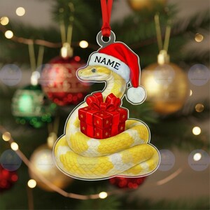 May include: A Christmas tree decoration featuring a yellow and white snake wearing a Santa hat with the word "NAME". The snake is coiled around a red gift box with a red bow. The ornament hangs from a red ribbon.