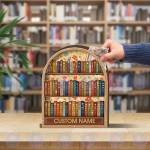 May include: A wooden bookshelf-shaped money bank with a floral interior and a row of colorful, decorative books. The bank has a slot at the top for inserting money. The text "CUSTOM NAME" is displayed on the bottom of the bank.