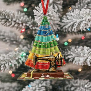 May include: A Christmas tree ornament featuring a sewing machine and a tree made of spools of thread in shades of green, blue, and yellow. The ornament has a red ribbon for hanging and the name "Lucie" is printed on the base.