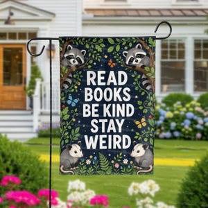 May include: A dark blue garden flag with the words "READ BOOKS BE KIND STAY WEIRD" in white. The flag features illustrations of raccoons, opossums, butterflies, and foliage. The flag is hanging on a black metal stand in a garden.