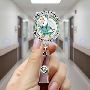 May include: A badge reel featuring a cartoon duck dressed as a doctor, holding a scalpel. The text on the badge reel reads "HERE'S THE THINGY WITH THE POINTY END." The reel has a clear plastic holder and a metal clip.