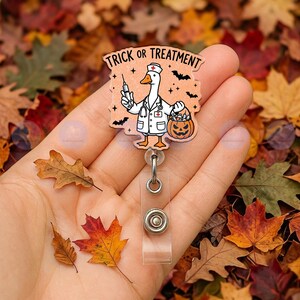May include: A badge reel featuring a peach-colored acrylic topper with a cartoon goose dressed as a nurse, holding a syringe and a jack-o'-lantern bucket. The text TRICK OR TREATMENT is printed at the top, accompanied by small black bats.