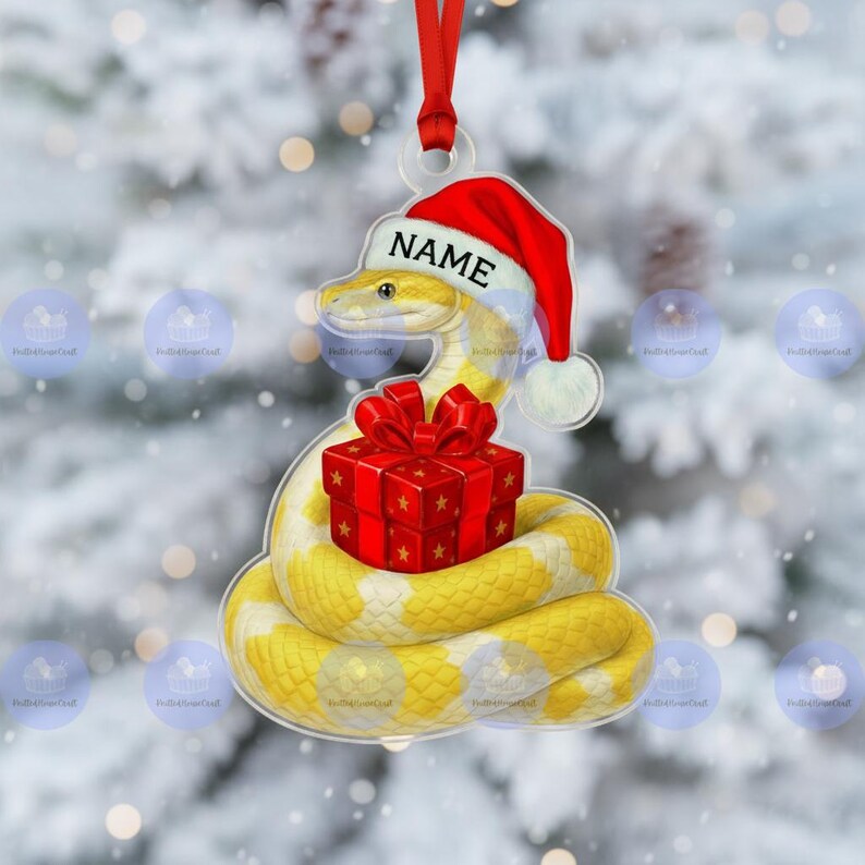 May include: A Christmas decoration showing a yellow and white snake wearing a Santa hat with the word "NAME". The snake is coiled around a red gift box with a red bow. The ornament hangs from a red ribbon.