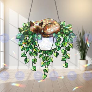 May include: Stained glass hanging art piece featuring a sleeping cat nestled in a white planter with cascading green leaves. The cat has a brown and tan mosaic pattern. The piece hangs from a silver chain.