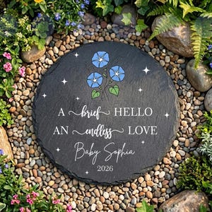 May include: A circular, dark grey slate garden marker inscribed with the words "A brief HELLO, AN endless LOVE, Baby Sophia, 2026." It has blue morning glory flowers and white star details. The stone is set amongst pebbles and plants.