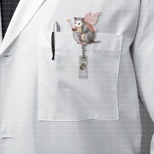 May include: A whimsical opossum badge reel with pink fairy wings and a golden wand. The opossum is gray and white, attached to a clear plastic reel with a metal clip. The badge reel is clipped to a white lab coat pocket.