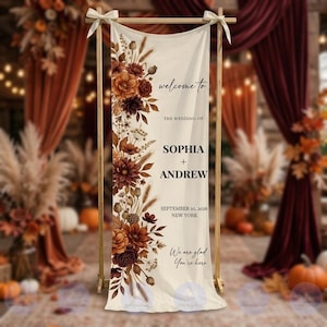 May include: A vertical cream-colored fabric wedding welcome sign suspended from a wooden frame. The left side features a floral arrangement in shades of rust, brown, and orange. Text reads: welcome to THE WEDDING OF SOPHIA + ANDREW SEPTEMBER 10, 2026 NEW YORK We are glad You're here.