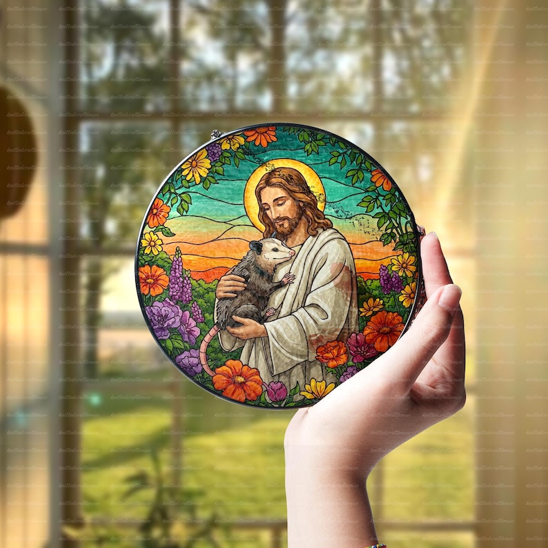 May include: A stained glass round ornament featuring a man with long hair and beard holding a small opossum. The man is wearing a long robe and the opossum is brown and white. The background is a colorful floral design.