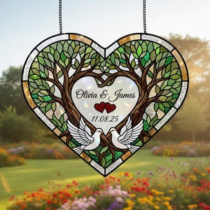Può includere: Vetrata a forma di cuore con un disegno ad albero, due colombe e i nomi "Olivia & James" con una data. La finestra presenta colori verdi, marroni e bianchi ed è sospesa da una catena.