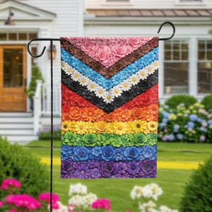May include: A garden flag featuring a vibrant floral design in a rainbow of colors. The flag showcases a chevron pattern with layers of flowers, including roses and daisies, creating a textured, dimensional effect. The flag is displayed on a black metal stand.
