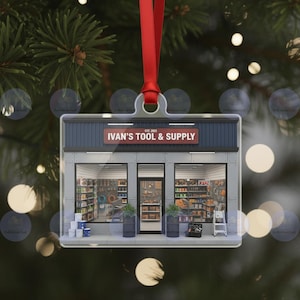 Könnte beinhalten: Ein klarer, rechteckiger Weihnachtsschmuck mit einer Miniatur-Fassade eines Baumarktes. Das Geschäft "Ivan's Tool & Supply" hat ein rotes Schild und ist mit Werkzeugen gefüllt. Der Schmuck hängt an einem roten Band vor einem verschwommenen Hintergrund aus Tannenzweigen und Lichtern.
