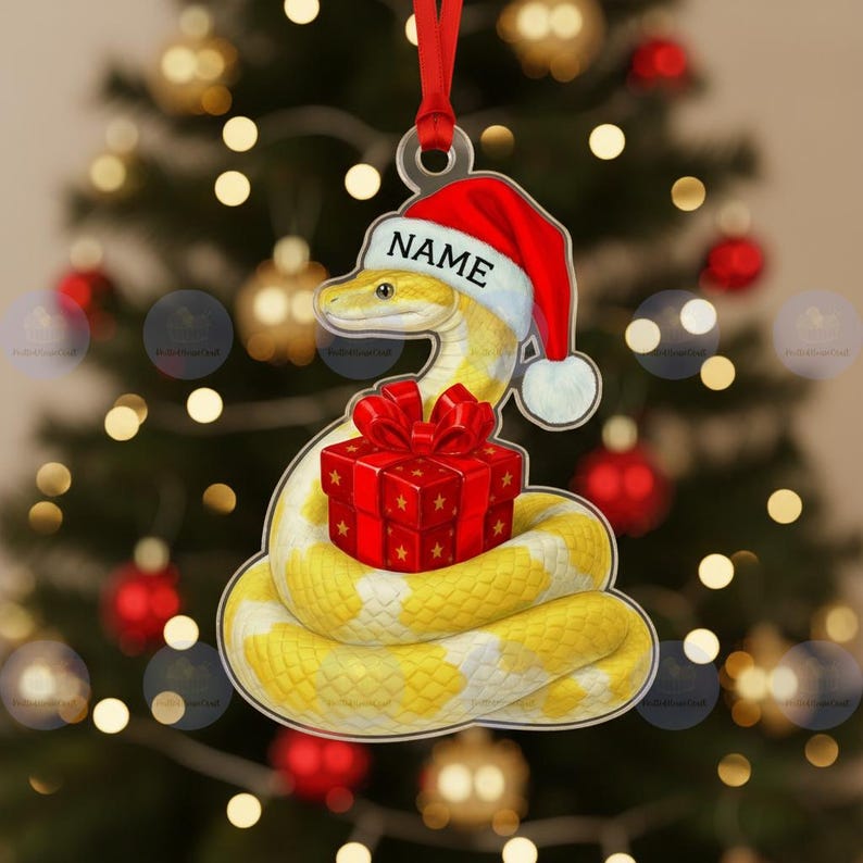 May include: A Christmas tree decoration depicting a yellow and white snake wearing a Santa hat with the word "NAME". The snake is coiled around a red gift box with a red bow. The ornament hangs from a red ribbon.