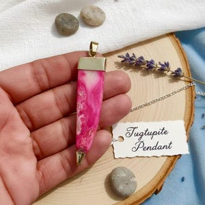 Puede incluir: Un colgante de tugtupita rosa y blanco con detalles metálicos dorados. El colgante es rectangular con un acabado pulido y está suspendido de una cadena plateada. La etiqueta dice "Tugtupite Pendant".