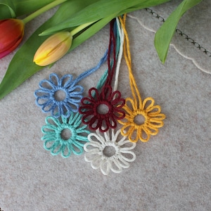 Könnte beinhalten: Sechs gehäkelte Blumenanhänger in verschiedenen Farben, darunter Blau, Türkis, Burgunder, Weiß und Gelb. Die Anhänger hängen an ihren Schnüren auf einer hellgrauen Oberfläche.