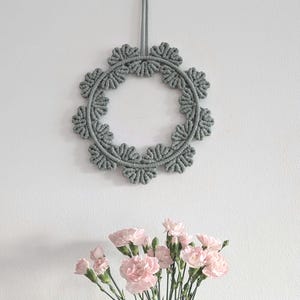 Könnte beinhalten: Ein grauer Makramee-Kranz hängt an einer weißen Wand mit einer Vase mit rosa Nelken davor.