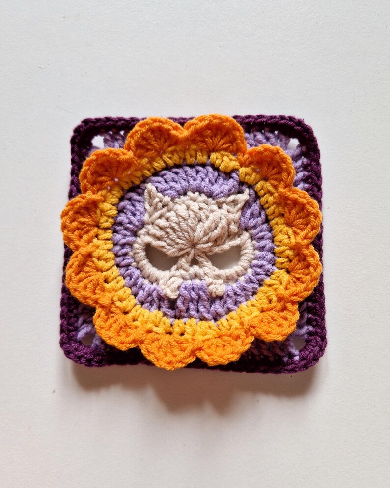 Vibrant version of the cat skull granny square using purples, yellows, and orange. The purple background and lavender details contrast beautifully with the skull and flower motif, making it a perfect choice for whimsical or gothic crochet projects.