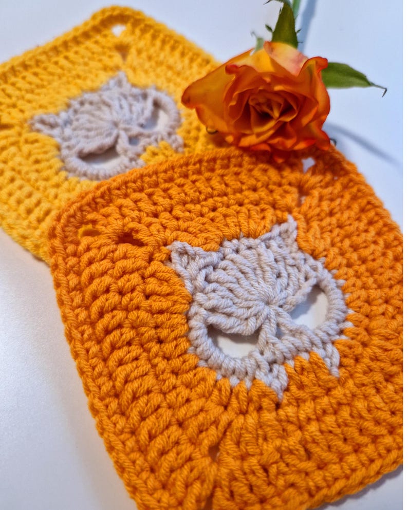 May include: Two crocheted squares, one yellow and one orange, each featuring a light gray owl design. A vibrant orange and yellow rose rests on the squares, adding a touch of natural beauty to the handmade crafts.