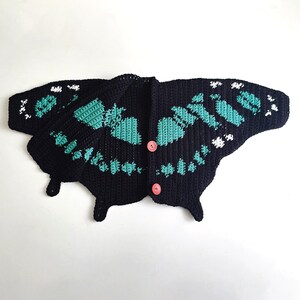 May include: A black crochet cardigan shaped like a butterfly, with turquoise and white accents. The butterfly cardigan has a button closure and is displayed on a white background. The wings are detailed with a turquoise pattern.