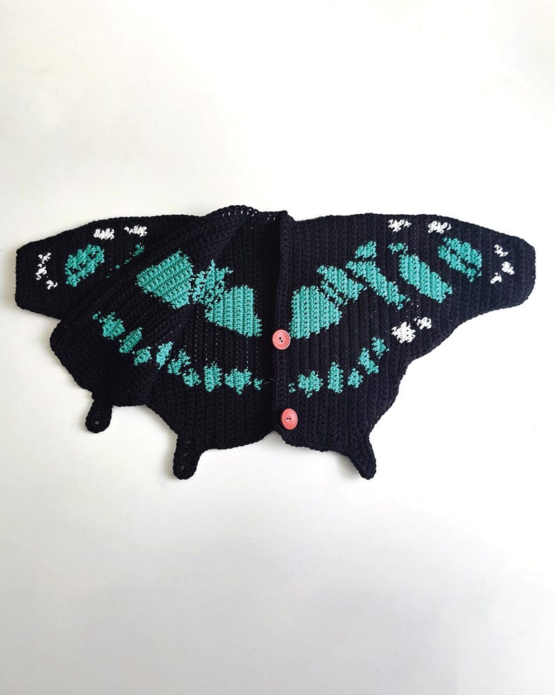 May include: A black crochet cardigan shaped like a butterfly, with turquoise and white accents. The butterfly cardigan has a button closure and is displayed on a white background. The wings are detailed with a turquoise pattern.