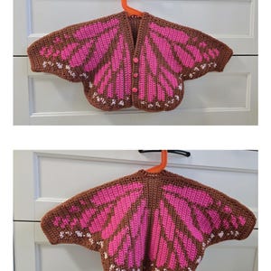 May include: A crochet cardigan featuring a brown and bright pink butterfly wing pattern with small white accents along the edges. The garment has a V-neckline, three pink buttons down the front, and wide, wing-shaped sleeves that extend outward.