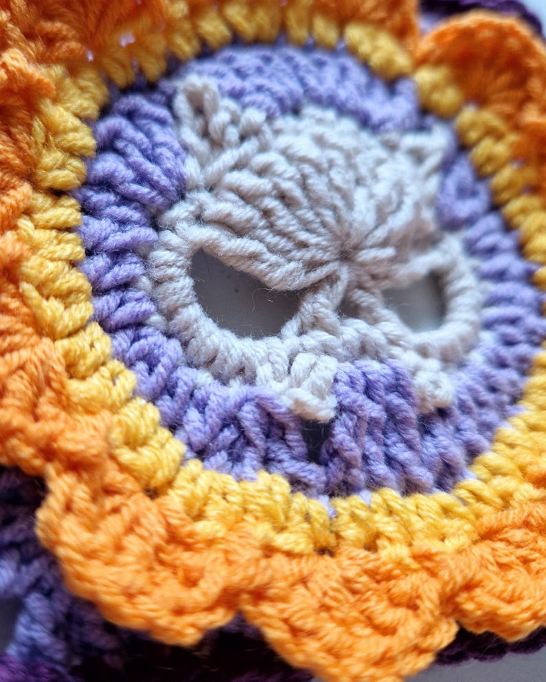 Peut inclure: Gros plan sur un motif de cr&acirc;ne crochet&eacute;. Le cr&acirc;ne est de couleur cr&egrave;me, entour&eacute; d'anneaux concentriques de fil violet, jaune et orange. Le travail au crochet est d&eacute;taill&eacute;, avec des points visibles cr&eacute;ant un effet textur&eacute;.