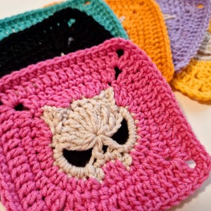 May include: A collection of colorful, crocheted squares. The front square is bright pink with a skull design in the center. Other squares are black, teal, orange, purple, and yellow. Each square has a square shape with a hole in each corner.
