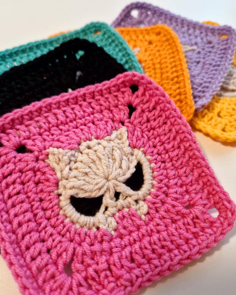 May include: A collection of colorful, crocheted squares. The front square is bright pink with a skull design in the center. Other squares are black, teal, orange, purple, and yellow. Each square has a square shape with a hole in each corner.
