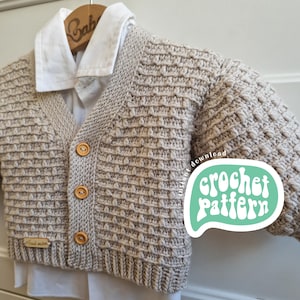 Può includere: Un cardigan beige lavorato a uncinetto con chiusura a bottoni. Il cardigan presenta un motivo a punto testurizzato ed è mostrato su un appendiabiti. Il testo "crochet pattern" è visibile nell'immagine.