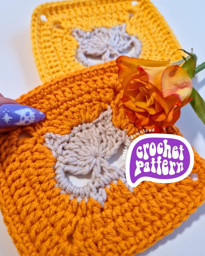 May include: Two crocheted squares in shades of yellow and orange, featuring a light beige fox design in the center. A vibrant orange rose and a purple sticker with the text "crochet pattern" are also visible.