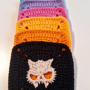 May include: A stack of colorful crochet squares. The squares are in shades of teal, purple, pink, yellow, orange, and black. The black square features a cream-colored skull design in the center. Each square has a square hole in each corner.
