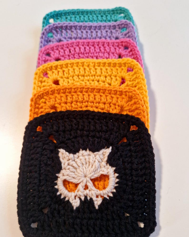 May include: A stack of colorful crochet squares. The squares are in shades of teal, purple, pink, yellow, orange, and black. The black square features a cream-colored skull design in the center. Each square has a square hole in each corner.