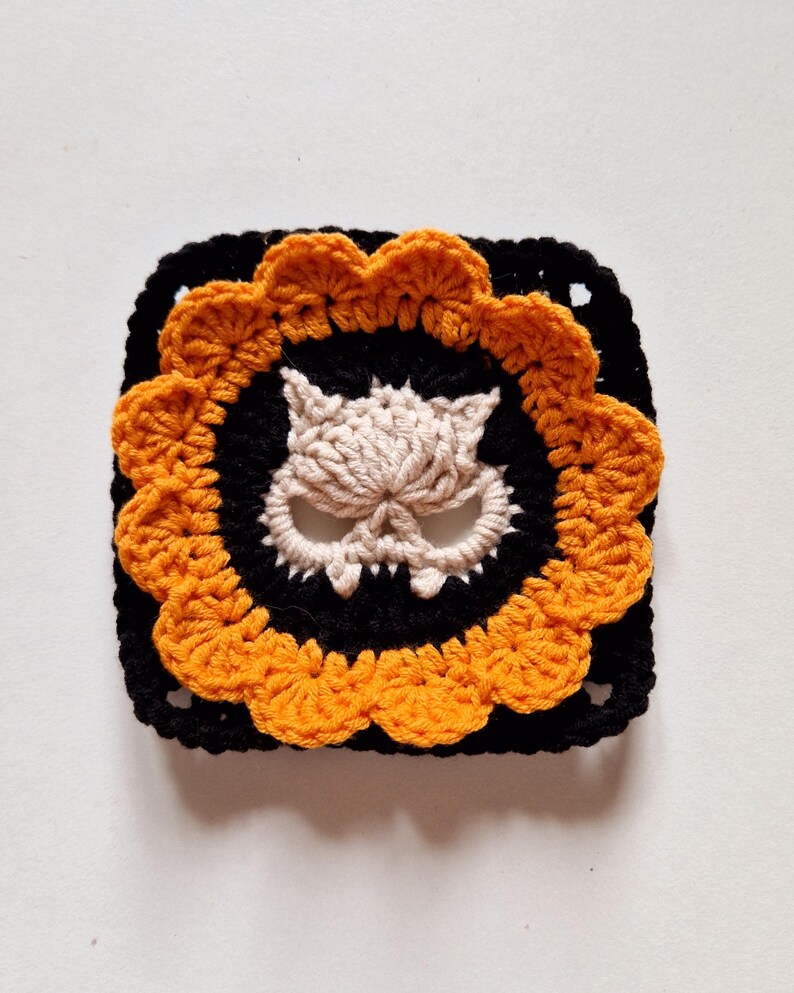 This crochet granny square features a classic Halloween color palette, 12 orange petals surrounding a cream cat skull motif on black background. The rich orange petals pop against the black square frame, making it perfect for Halloween bunting, table runners, or spooky blankets. For crafters who love alternative designs, punk or goth crochet aesthetics, or want to add a unique twist to their Halloween handmade creations.